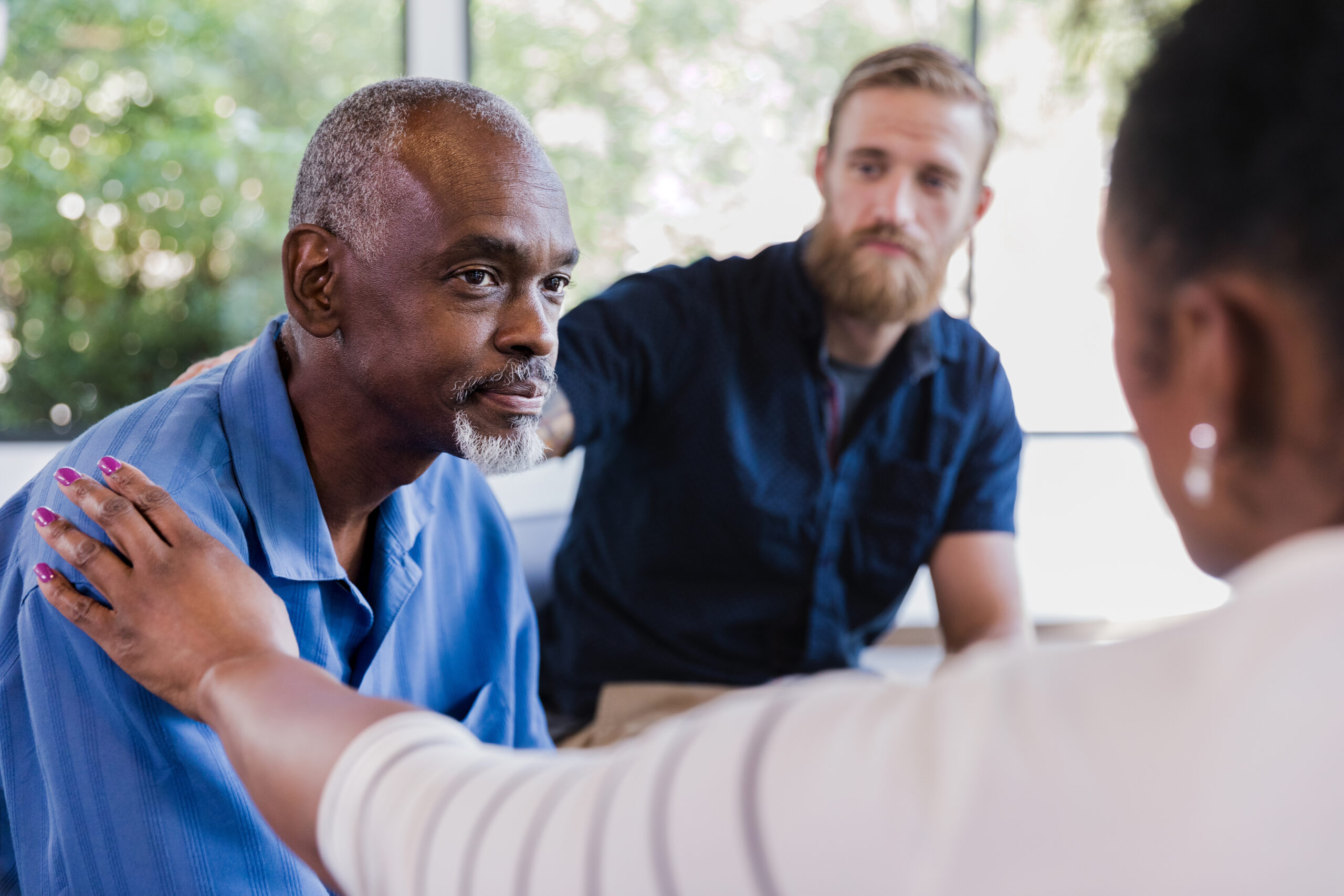 Mature man attentively listens as a female therapist comforts and encourages him during a group therapy meeting. The therapist's hand is on the man's shoulder.